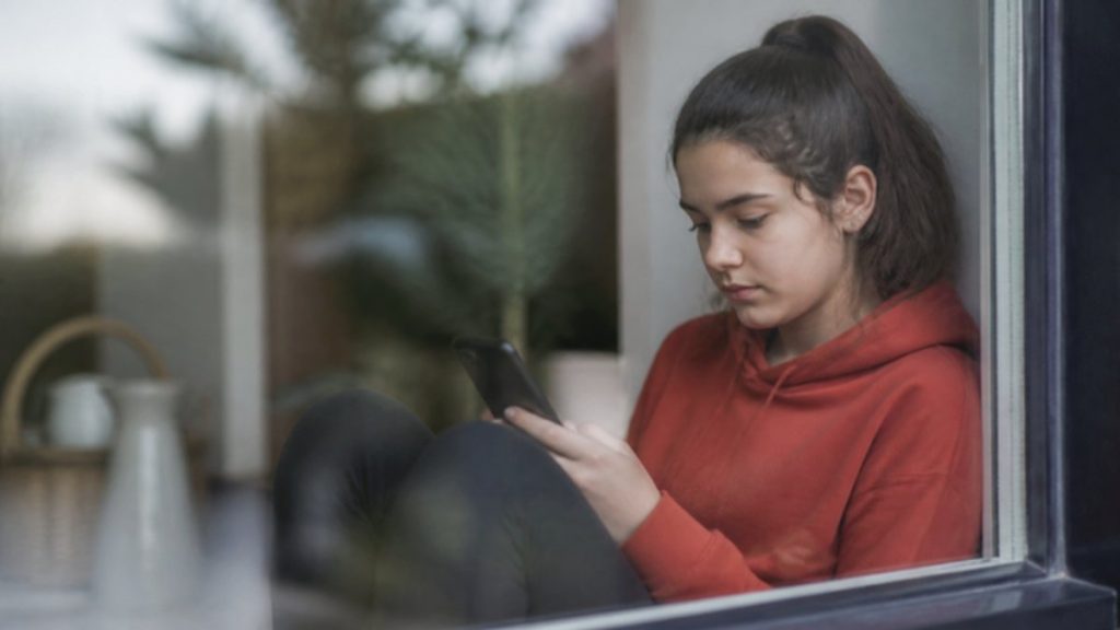 Adolescente sola junto a la ventana conversando con un chatbot de IA en el móvil